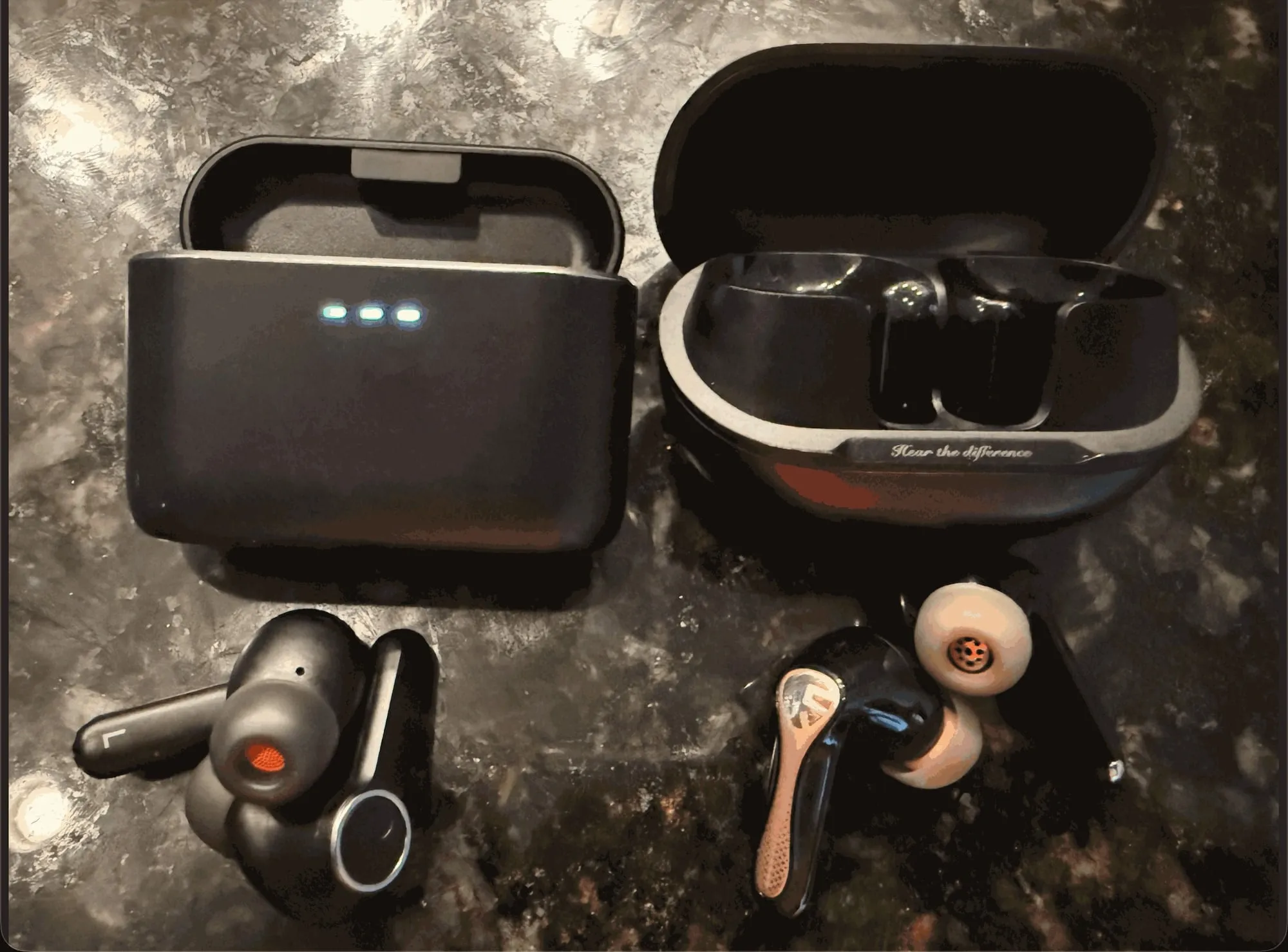 The Cambridge Audio Melomania A100 (left) and the Soundpeats Air5 Pro+ (right) on a dark stone counter, each with its open case behind and its buds in front; the Air5 Pro+ buds have gold stems.