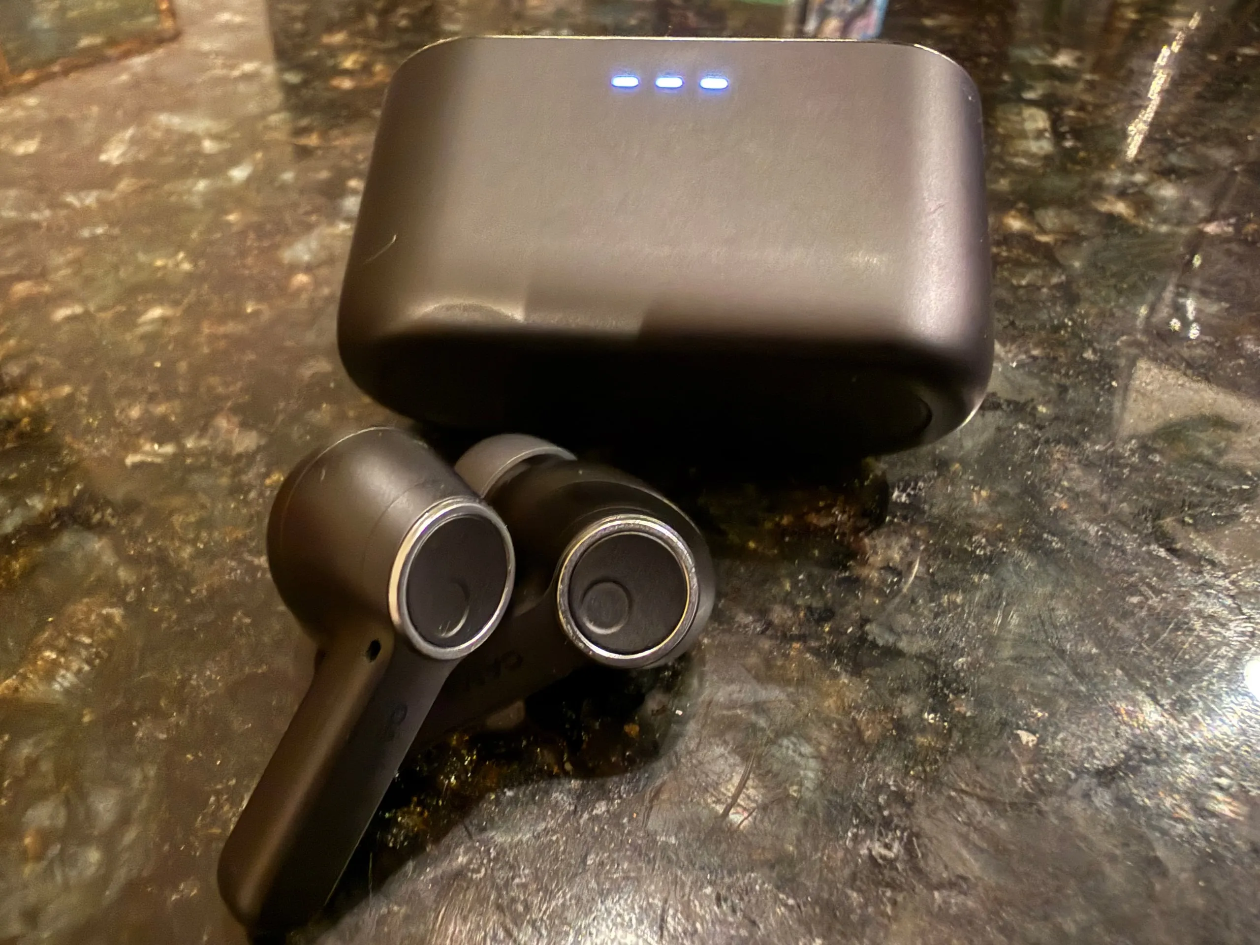 The Cambridge Audio Melomania A100 buds lying on a dark stone counter in front of their closed case, its three charging lights lit; the buds show their round, metal-rimmed touch surfaces.