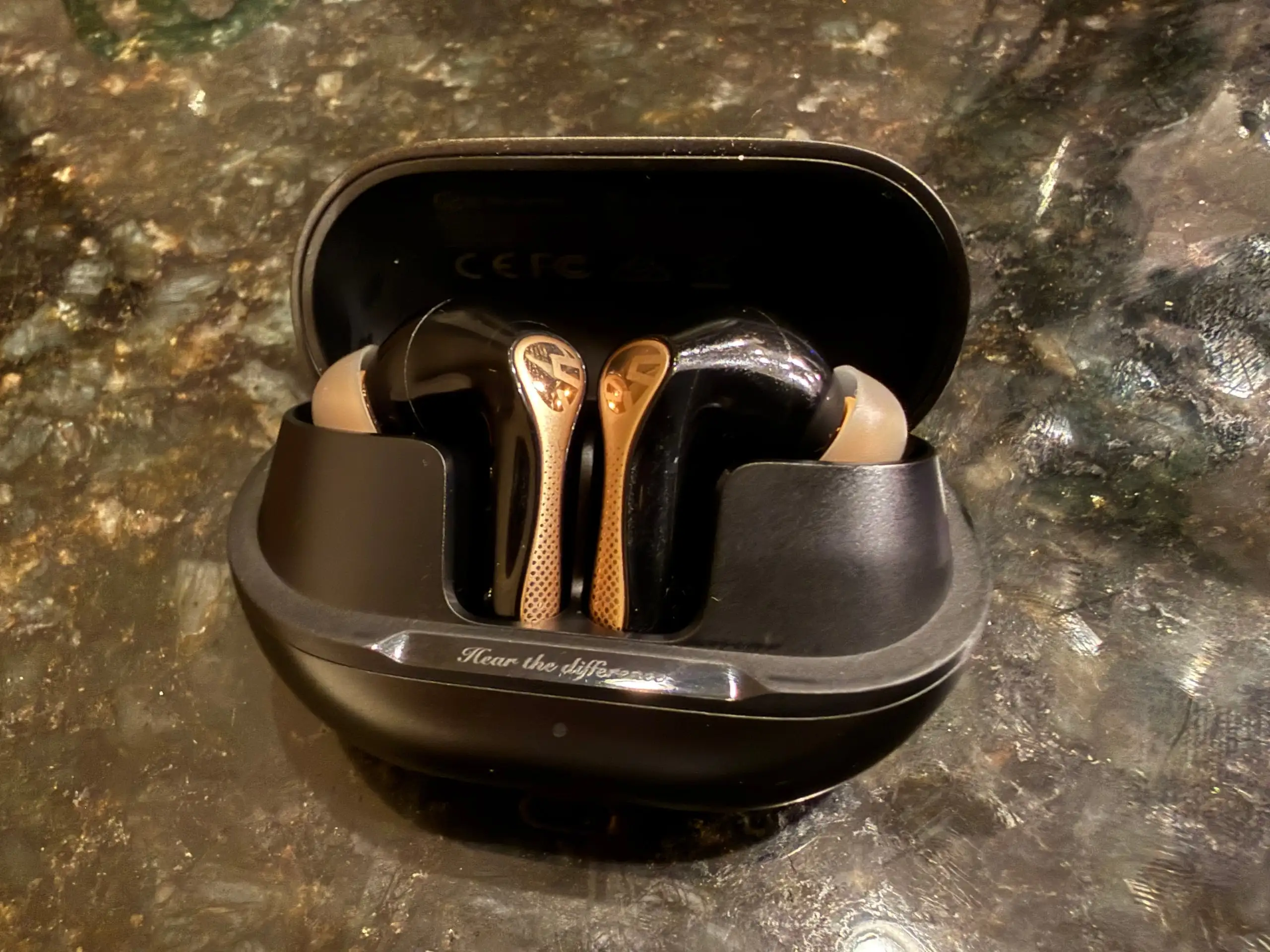 The Soundpeats Air5 Pro+ in its open black case on a dark stone counter, glossy black buds with perforated gold stems.