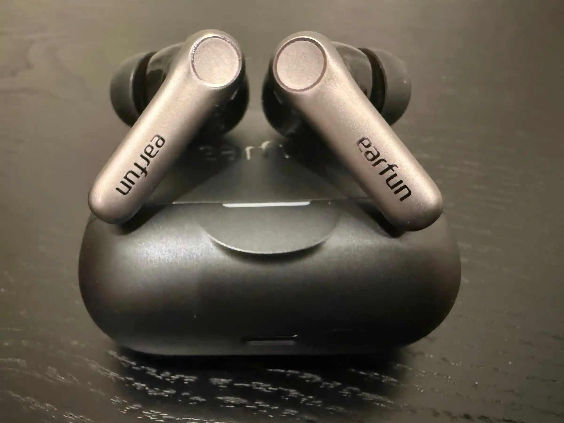 Both EarFun Air Pro 4+ buds resting on top of their closed black case on a dark wooden table, their brushed grey stems printed with the earfun name and their round touch pads facing up.