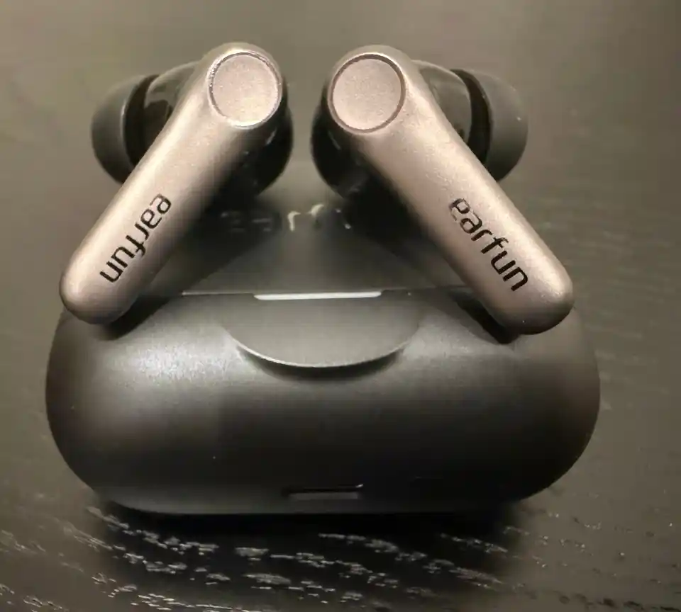 Both EarFun Air Pro 4+ buds resting on top of their closed black case on a dark wooden table, their brushed grey stems printed with the earfun name and their round touch pads facing up.