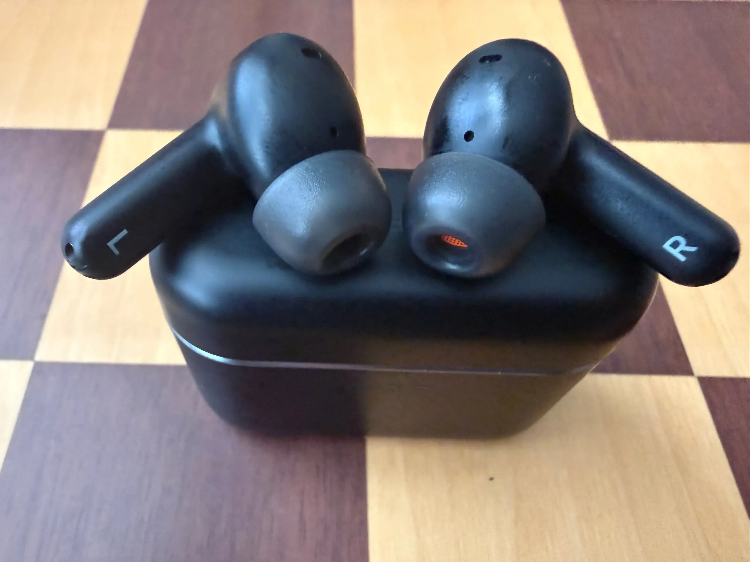 Both Melomania A100 buds resting on top of their closed case on a chessboard, stems marked L and R, the right nozzle showing its orange mesh.