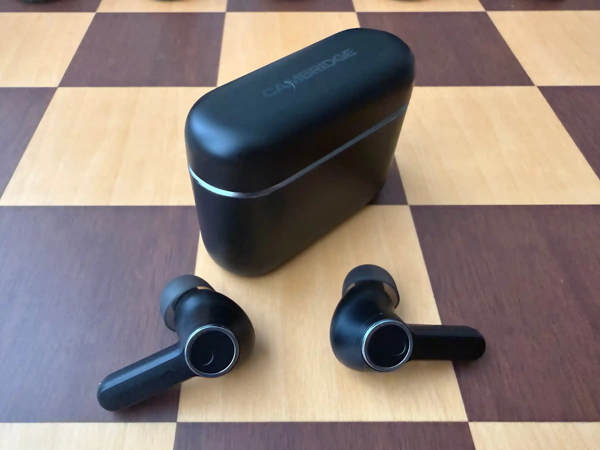 The closed Melomania A100 case in matte black with a thin metal band, on a chessboard, and both buds in front of it showing their round touch surfaces ringed in metal.