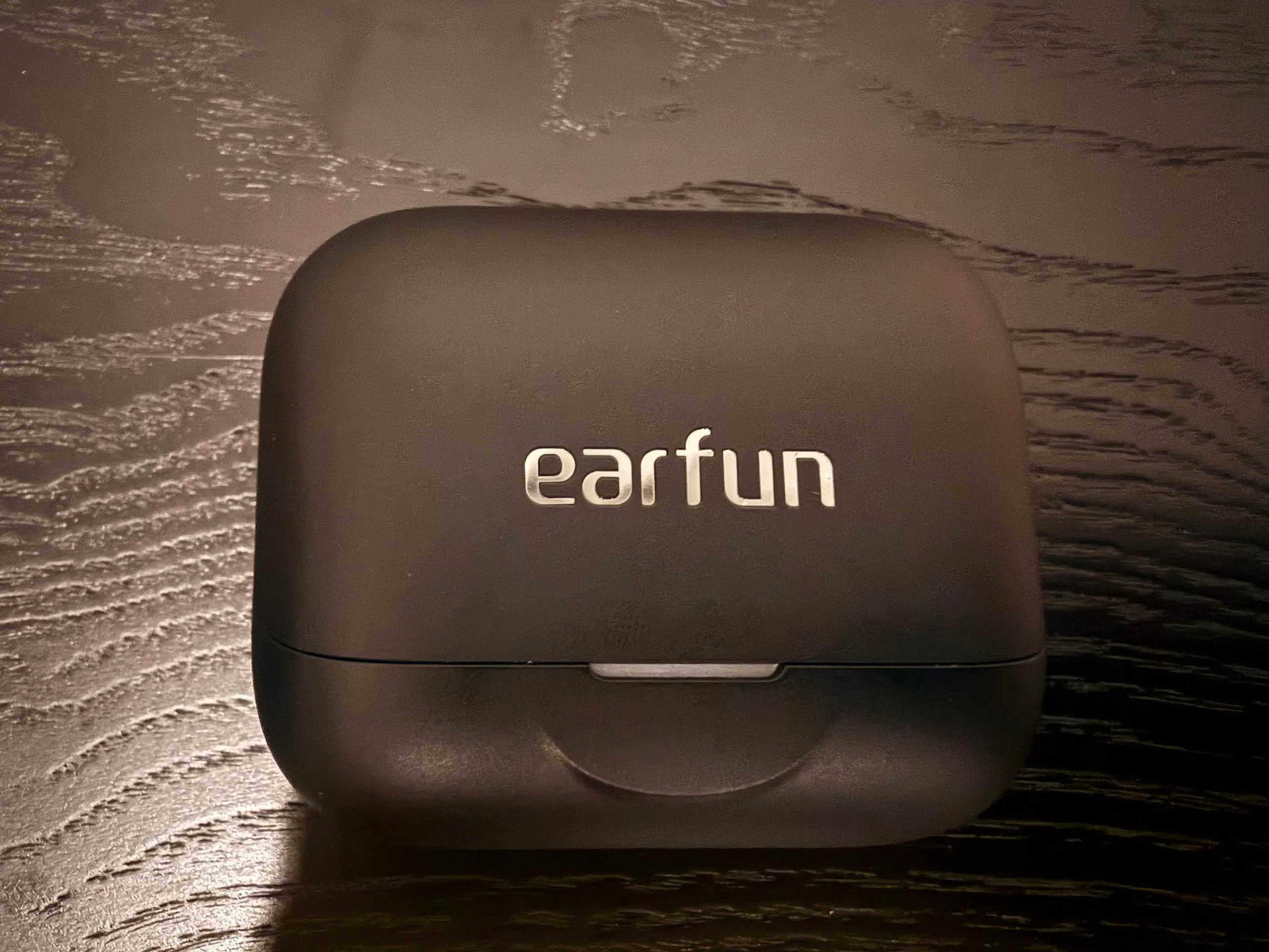 The closed EarFun Air Pro 4+ case on a dark wooden table, matte black with the earfun name in silver on the lid, a small status light and a curved cut-out on the front.