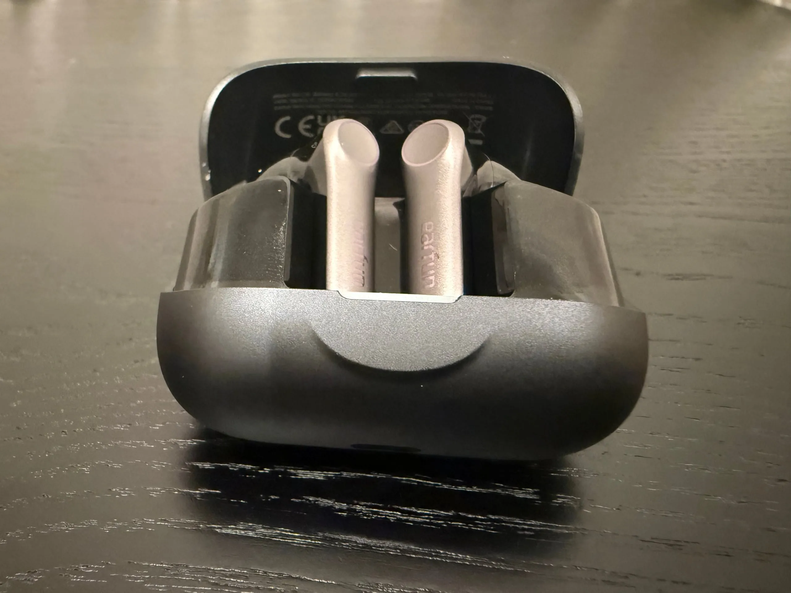 The EarFun Air Pro 4+ case opened from the front on a dark wooden table, both grey stems standing in a clear inner tray, the lid's regulatory markings behind them.