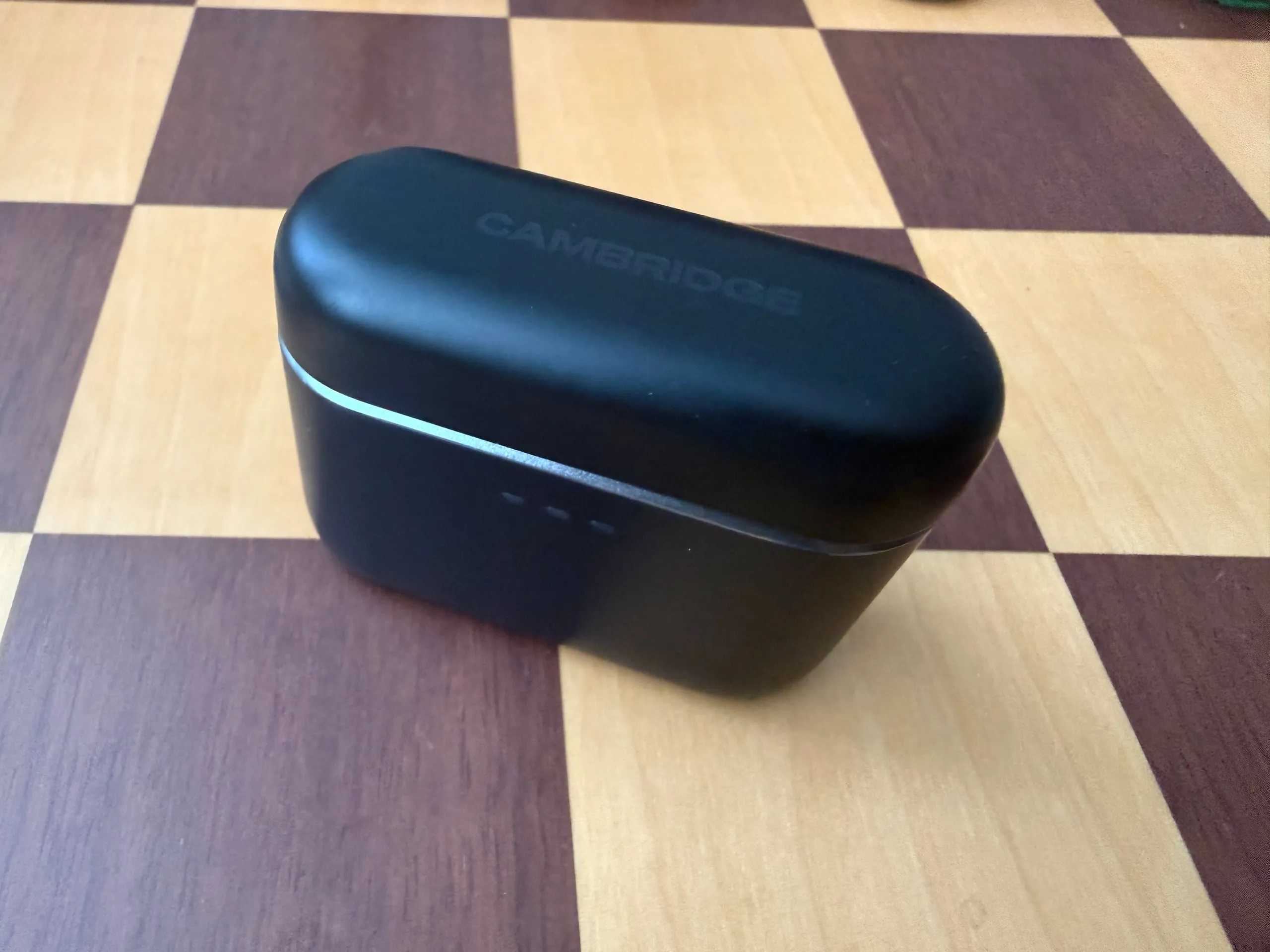 The closed Melomania A100 case on a chessboard: matte black with CAMBRIDGE printed on the lid, a thin metal band and three small charging lights on the front.