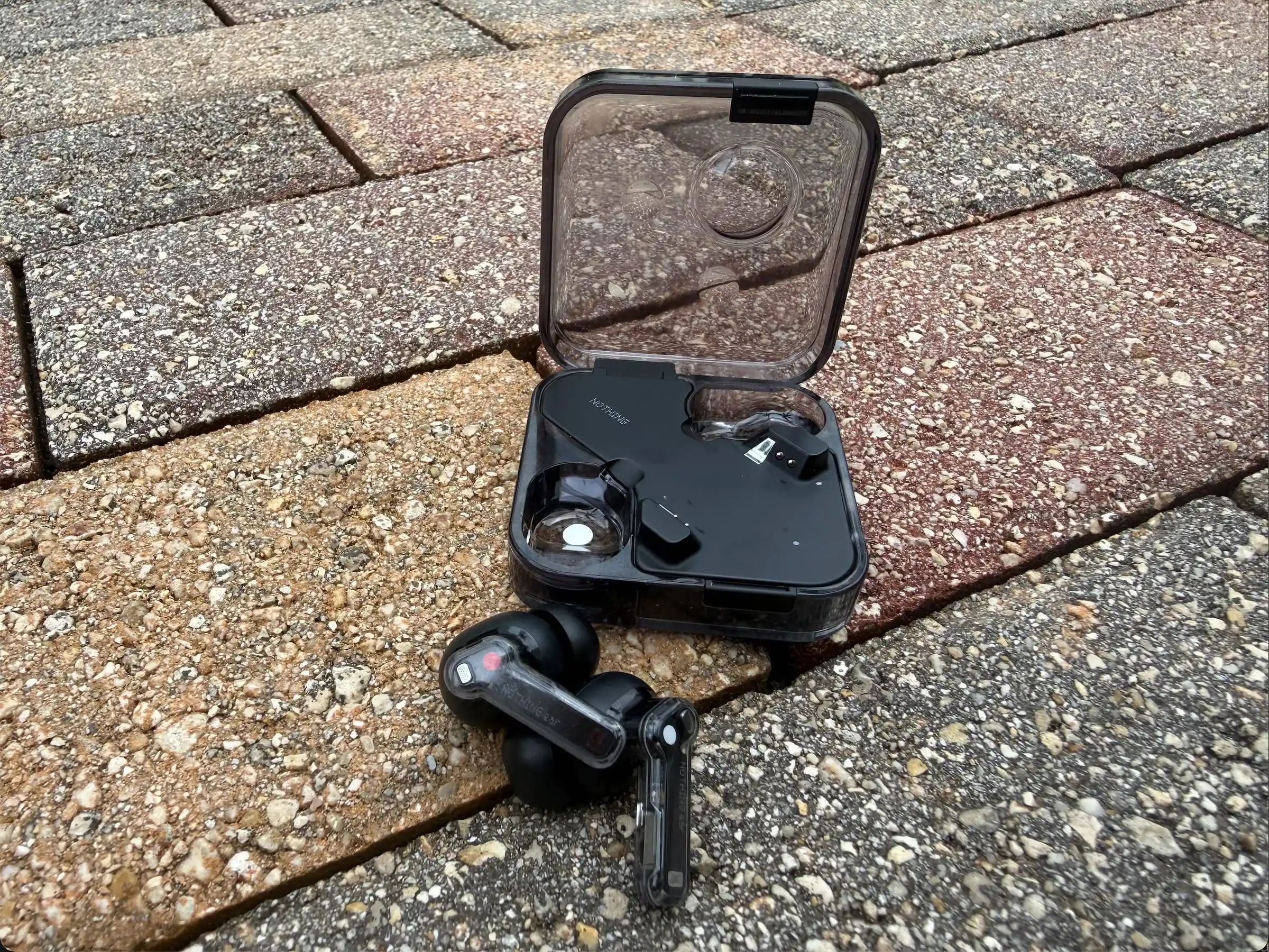 The Nothing Ear (2024) buds lying on brick paving in front of their open transparent case.