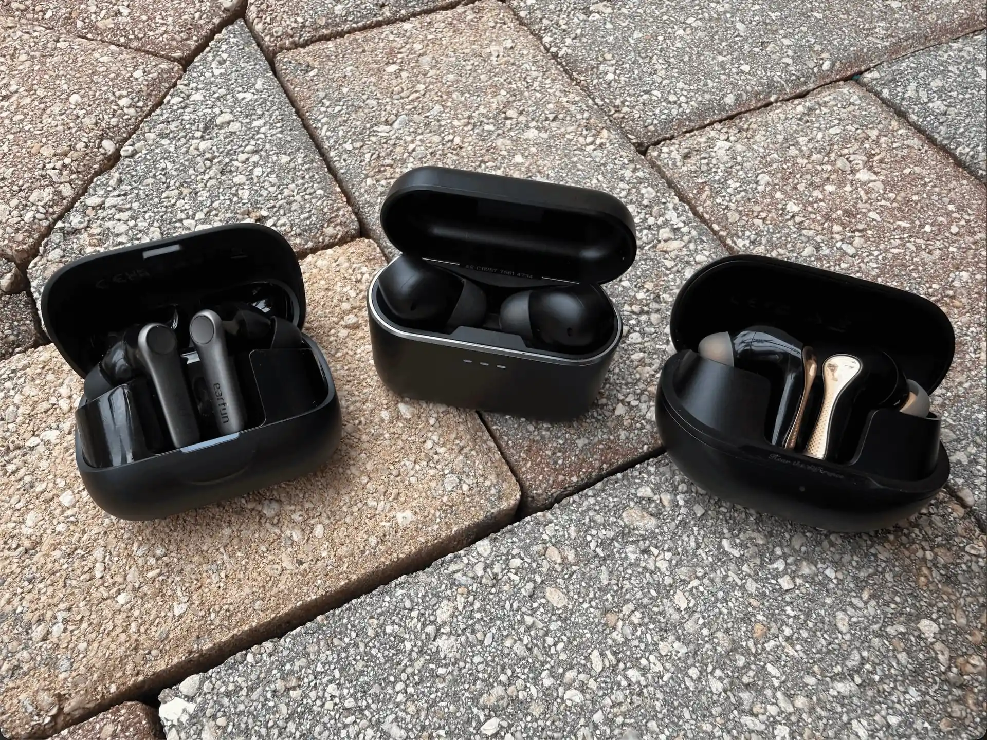 Three open charging cases on brick paving: the EarFun Air Pro 4+ on the left, the Cambridge Audio Melomania A100 in the middle, and the Soundpeats Air5 Pro+ with its gold stems on the right.